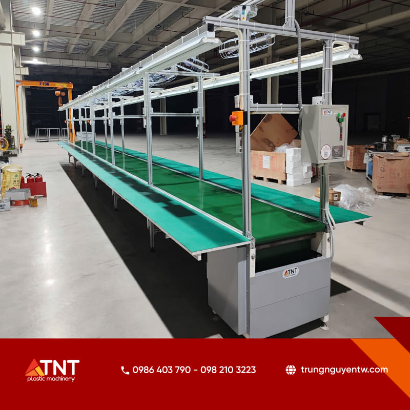 Hệ thống băng tải PVC dài kết hợp dàn đèn chiếu sáng và bàn thao tác hai bên trong nhà xưởng công nghiệp